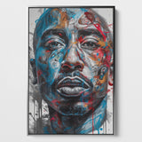 Das Bild zeigt ein lebendiges Porträt von Tupac Shakur mit expressiven, farbenfrohen Akzenten auf einer Leinwand. Der Stil des Kunstwerks ist modern und wirkt fast wie eine Fusion aus Realismus und abstrakter Kunst. Die Farben Blau und Rot dominieren, wobei dynamische Farbspritzer und Ströme über das Gesicht laufen, was dem Bild Energie und Bewegung verleiht. Das Bild ist eingerahmt in einem silbernen Schattenfugenrahmen.